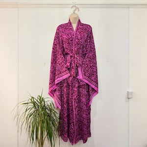 May include: A magenta-colored robe with a floral pattern, hanging on a mannequin. The robe has a matching belt and trim. The floral design is a darker shade of magenta. The robe is displayed against a white wall with a green plant in the foreground.
