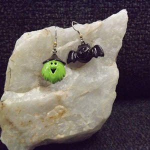May include: A pair of Halloween-themed earrings. One earring features a green witch with a black hat and a smiling face. The other earring features a black bat with wings.