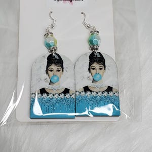 May include: A pair of dangle earrings featuring an image of a woman with a bubblegum bubble. The earrings have silver hooks and are adorned with blue and green beads. The image is set against a white background.