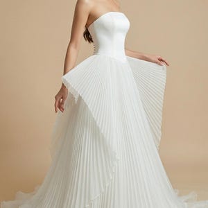May include: A white strapless wedding dress with a pleated skirt and a flowing, layered design. The bodice is fitted, and the skirt flares out, creating a dramatic silhouette. The dress is made of a soft, flowing fabric.