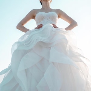 May include: A strapless white wedding dress with a layered skirt and a lace bodice. The dress has a flowing, voluminous design. The image is taken from a low angle, looking up at the dress against a clear, light blue sky.