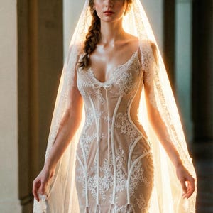 May include: A bridal gown featuring a fitted bodice with intricate white lace detailing over a nude underlay. The dress has a V-neckline and a flowing skirt. A sheer veil drapes from the head, completing the elegant ensemble.
