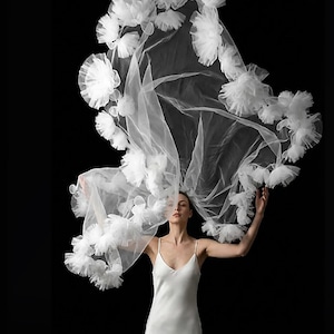 Puede incluir: Un velo de novia blanco con detalles florales se muestra sobre un fondo negro. El velo está adornado con numerosas decoraciones blancas en forma de flor. La modelo lleva un vestido blanco con tirantes finos.