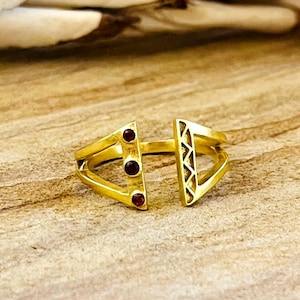 May include: A gold-toned open ring featuring a geometric design. One side of the band displays three small, round, dark red stones set vertically, while the opposite side features a triangular cutout with a textured, zigzag pattern engraved inside.