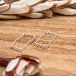 Puede incluir: Un par de pendientes de aro rectangulares plateados. Los pendientes se muestran sobre una superficie de madera con vetas naturales. El fondo incluye vainas de semillas secas y una flor parcialmente visible.