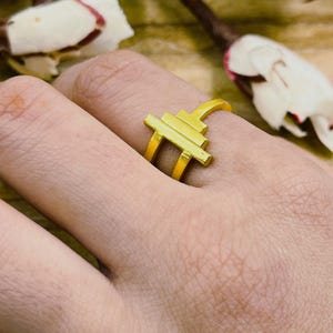 May include: A gold ring with a geometric design. The ring features a series of horizontal and vertical bars, creating a modern and abstract look. The ring is worn on a finger, showcasing its bright, shiny finish. The ring's design is simple yet eye-catching.