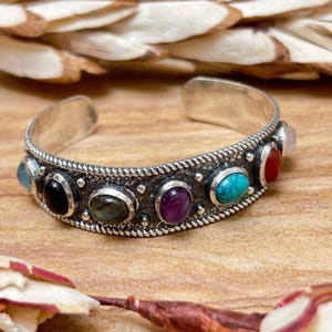 May include: A silver cuff bracelet with an open design, featuring a row of colorful gemstones. The bracelet has a textured border and is adorned with oval and round stones in various colors, including turquoise, red, and purple. The bracelet is displayed on a wooden surface.