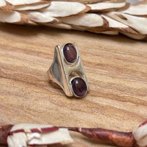 May include: A silver ring with two oval, dark purple gemstones. The ring has a geometric design with a rectangular shape and a split band. The gemstones are set in a bezel setting. The ring is displayed on a wooden surface.