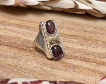 May include: A silver ring with two oval, dark purple gemstones. The ring has a geometric design with a rectangular shape and a split band. The gemstones are set in a bezel setting. The ring is displayed on a wooden surface.