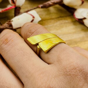 Può includere: Un anello dorato dal design unico. L'anello presenta due elementi sovrapposti a forma di foglia. L'anello è indossato su un dito, mostrando la sua finitura metallica brillante. Il design dell'anello è moderno e accattivante.