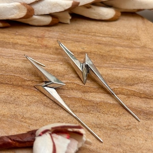 May include: A pair of silver lightning bolt earrings. The earrings have a geometric design with sharp angles and a long, pointed bottom. The earrings are displayed on a light brown surface.