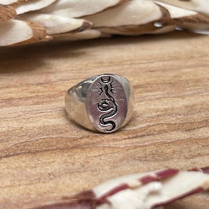 May include: Silver ring with an oval face featuring a detailed black snake design. The snake is coiled and positioned beneath a crescent moon and sunburst motif. The ring has a polished finish and a thick band.