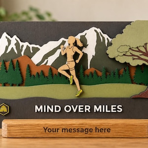 May include: A layered wooden sign featuring a runner in the center, a mountain range, pine trees, and a large tree. The text "MIND OVER MILES" is printed in white on a dark base. A wooden stand at the bottom includes the text "Your message here".