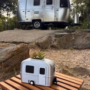 May include: A silver-colored, miniature travel trailer planter featuring a succulent plant growing from the roof. The planter is shaped like a classic rounded camper with black window and door details, resting on a slatted wooden surface outdoors.