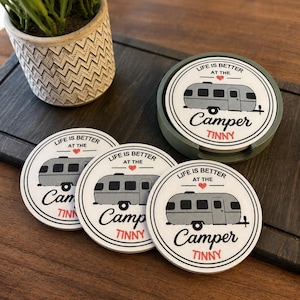 May include: A set of four round white coasters with a gray camper illustration, a red heart, and the black text "LIFE IS BETTER AT THE Camper" with the red word "TINNY" underneath. One coaster is stored inside a circular olive green holder.