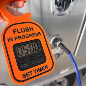 May include: A bright orange plastic hook-shaped timer with a digital display showing 05:00. The text FLUSH IN PROGRESS is printed in black above the screen, and SET TIMER is printed below. The device features three black buttons for control.
