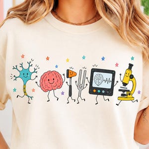 May include: A cream-colored t-shirt featuring a graphic print of cartoon-style medical and scientific icons including a neuron, a brain, a reflex hammer, a tuning fork, a brain scan monitor, and a microscope, all accented with small colorful stars.