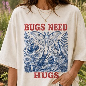 May include: An off-white t-shirt featuring a blue graphic illustration of various insects including a moth, bee, praying mantis, beetle, ladybug, and caterpillar among plants and mushrooms, with the red text BUGS NEED HUGS printed above and below.