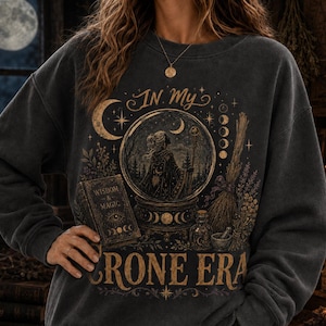 May include: A charcoal gray crewneck sweatshirt featuring a gold-toned graphic print. The design includes a crystal ball with a figure, a book titled Wisdom & Magic, a broom, and the text In My Crone Era, surrounded by moon phases and stars.