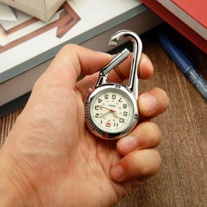 Puede incluir: Un reloj de enfermera plateado con esfera blanca y números negros. El reloj tiene un segundero rojo y un mosquetón para sujetarlo. El reloj se sostiene en una mano.