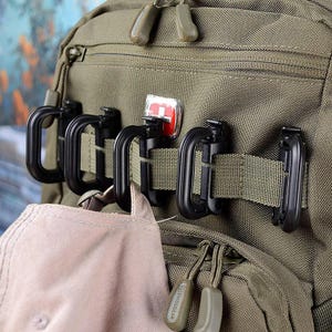 May include: Close-up of a green backpack with black carabiner clips attached to the exterior. The clips are secured with olive green webbing. A red and white logo is visible. A tan baseball cap is partially visible.