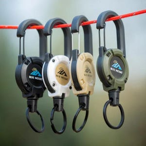 May include: Four retractable keychains in black, white, tan, and olive green hang from a red rope. Each keychain has a carabiner clip and a black key ring. The keychains have the text "BUYE MOUNT" and a mountain logo.
