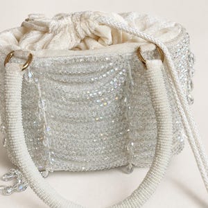 May include: A white drawstring handbag covered in clear, faceted beads that create a shimmering texture. The bag features two rounded, fabric-covered handles attached with gold-tone rings and a soft, gathered fabric interior closure.
