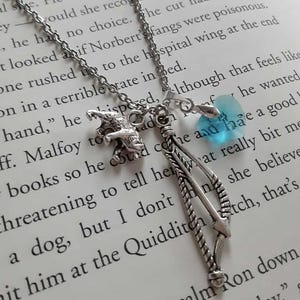 May include: Silver necklace with charms including a dragon, a bow and arrow, and a blue heart-shaped crystal. The necklace is shown on a book with printed text.