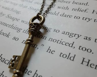 May include: A bronze-coloured key pendant necklace. The key, with detailed features, hangs from a matching chain. The necklace is set against a backdrop of book pages, creating a vintage aesthetic.