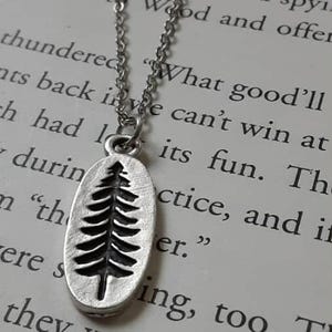 Peut inclure: Un collier argenté avec un pendentif ovale représentant un arbre. Le pendentif est suspendu à une fine chaîne. L'arrière-plan est une page de texte imprimé.
