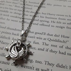 May include: A silver-toned necklace featuring a pendant with a swan inside a ship's wheel design. The necklace is displayed on a book page with printed text. The swan pendant is a detailed, decorative piece.