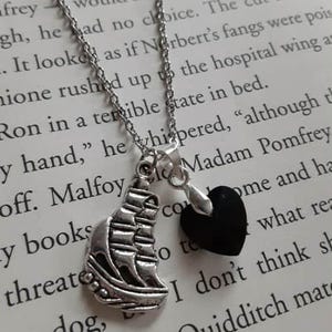 Könnte beinhalten: Halskette mit silberfarbener Kette und zwei Anhängern: ein detailliertes Segelboot und ein schwarzes Herz. Die Kette liegt auf einer Buchseite mit gedrucktem Text.
