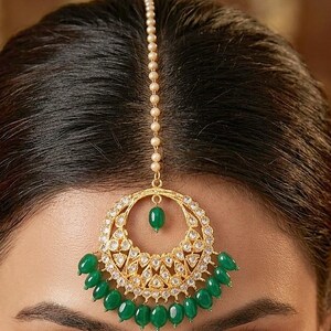 May include: A gold-toned headpiece with a circular pendant and a vertical chain of small beads. The pendant is adorned with clear crystals and hanging green beads. The headpiece is worn on the forehead.