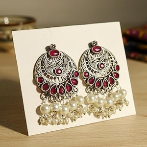May include: Silver-coloured earrings with an intricate design. Each earring has a central red gemstone, surrounded by smaller red gems and dangling white pearls. The earrings are presented on a white card.