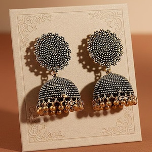 Puede incluir: Un par de pendientes jhumka ornamentados, de color plata oscura, con un ribete dorado y pequeñas cuentas colgantes. Los pendientes se exhiben en una tarjeta color crema con un diseño floral, sobre un fondo marrón cálido.