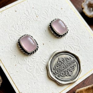 Op de afbeelding: Vierkante rozenkwarts oorknopjes met een gedraaide zilveren rand. De oorbellen worden gepresenteerd op een decoratieve kaart, samen met een zilveren zegel met de tekst "TAJUNCREATIONS".