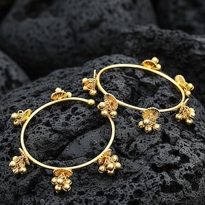 Puede incluir: Dos brazaletes dorados con detalles florales y de cuentas. Los brazaletes circulares descansan sobre una superficie oscura y texturizada. Cada brazalete presenta múltiples elementos decorativos, incluyendo pequeños grupos de cuentas y diseños florales estilizados.