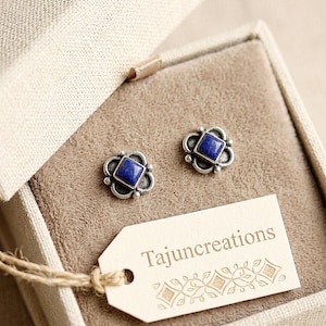 May include: A pair of silver-toned stud earrings with square, deep blue centers, displayed in a beige jewelry box. The earrings are accented with a floral design. A tag with the text "Tajuncreations" is attached.