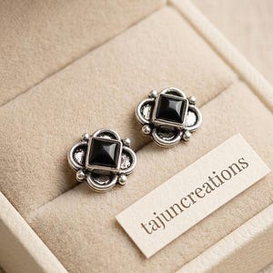 May include: Two silver-coloured stud earrings, each featuring a square black stone at the centre, set within a decorative floral design. The earrings are presented in a beige jewellery box. A tag with the text "tajuncreations" is also visible.