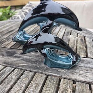 Peut inclure: Deux sculptures de dauphins en céramique. Les dauphins noirs ont une finition brillante, sautant au-dessus de vagues bleues. Les sculptures sont exposées sur une table en bois vieilli, mettant en valeur un thème nautique.