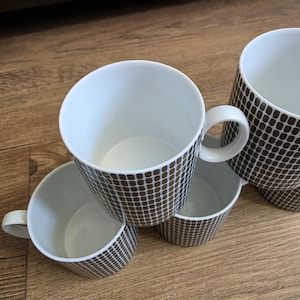 Pode incluir: Um conjunto de três canecas de cerâmica branca com um padrão de grade castanho-escuro repetido no exterior. Cada caneca tem um interior branco liso, uma pega arredondada e uma forma cilíndrica. As canecas estão dispostas em grupo.