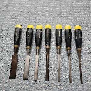 May include: A set of seven wood chisels arranged in a row, each featuring a black handle with a yellow cap and a metallic blade. The handles are marked with the brand name Footprint, and the blades vary in width from wide to narrow.