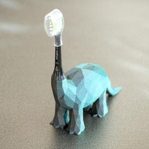 May include: A teal and gray geometric dinosaur-shaped toothbrush holder with a black toothbrush. The toothbrush head is covered with a clear plastic cap. The holder is on a gray surface.