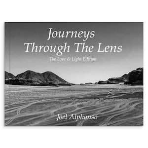 May include: A black and white book cover with the title "Journeys Through The Lens" and the subtitle "The Love & Light Edition." The cover features a landscape photo of a desert with mountains and tire tracks. The author's name, Joel Alphonso, is at the bottom.