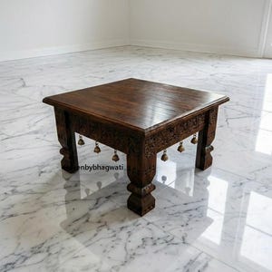 May include: A square, dark brown wooden table with carved floral designs along the edges and decorative bells hanging from the bottom. The table has a polished surface and stands on four ornate legs. The table is approximately 24 inches wide and 18 inches tall.