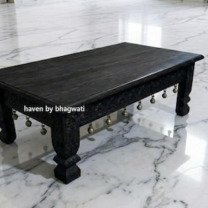 Könnte beinhalten: Ein rechteckiger, schwarzer Couchtisch aus Holz mit geschnitzten Beinen und einem dekorativen Rand. Kleine, goldfarbene Glöckchen hängen am Rand des Tisches. Der Tisch ist ca. 91 cm lang, 61 cm breit und 30 cm hoch. Der Tisch steht auf einem Marmorboden.