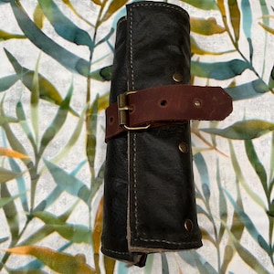May include: A rolled black leather case, fastened with a brown leather strap and a brass buckle. The case is stitched with light thread and has brass rivets. The background features a watercolour botanical print with green, yellow, and blue leaves.