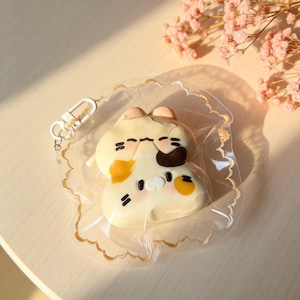 May include: A cream-coloured cat-shaped keyring with black and orange accents, encased in a clear plastic pouch with a gold trim. The keyring has a silver clasp. The background is a light-coloured surface with pink flowers.