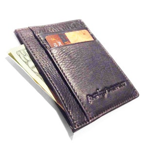 May include: A dark brown leather cardholder wallet with a textured finish. The wallet has multiple slots, one of which holds a credit card. A twenty-dollar bill is partially visible. The brand name is embossed on the front.