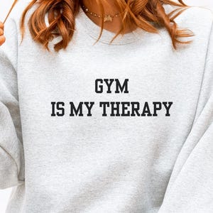 May include: A light gray sweatshirt with the text "GYM IS MY THERAPY" in black. The sweatshirt is paired with light blue jeans. The person is sitting on a yellow and white checkered surface.